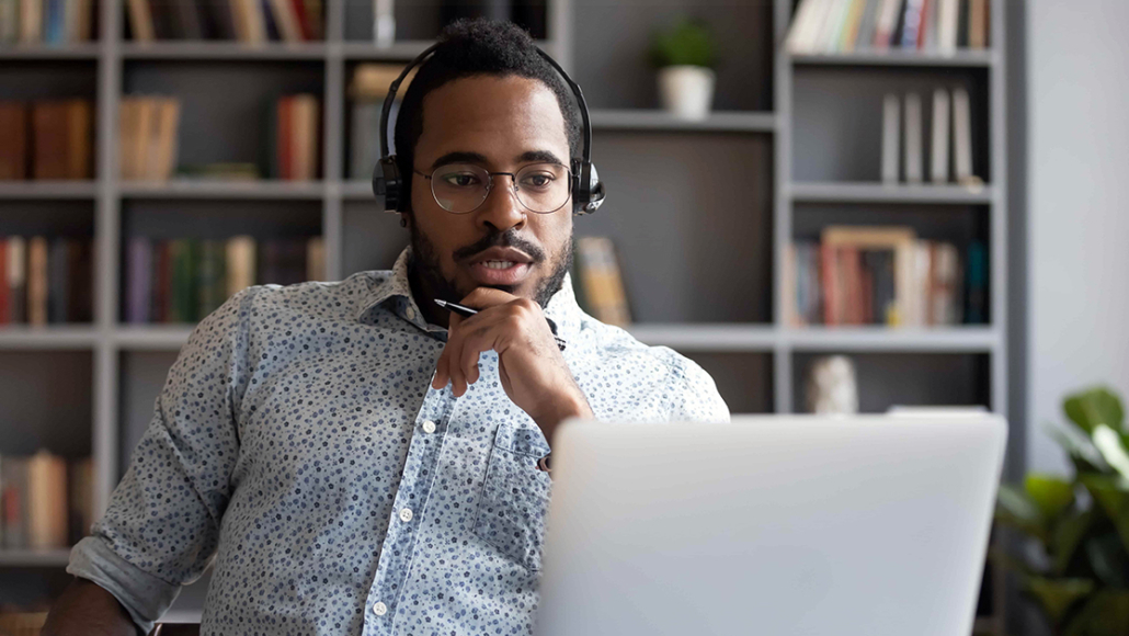 A professional man wearing glasses and headphones sits at a desk, engaged in an online meeting. He appears focused while looking at a laptop, with bookshelves and plants in the background.
